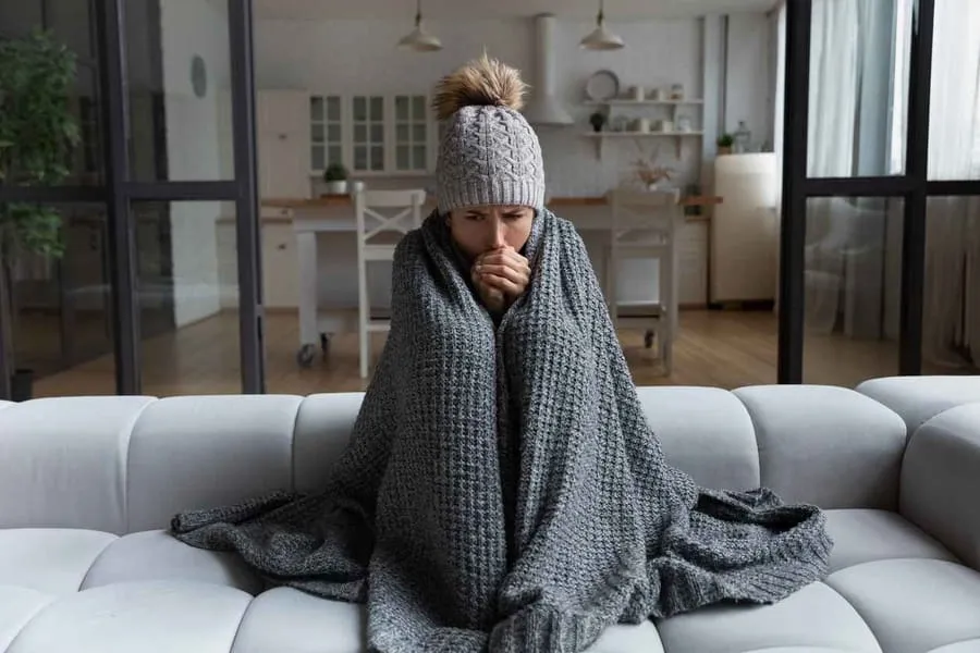Woman sitting on couch with a blanket wrapped her because her heater is broken. She is wearing a hat and covering her mouth with her hands.